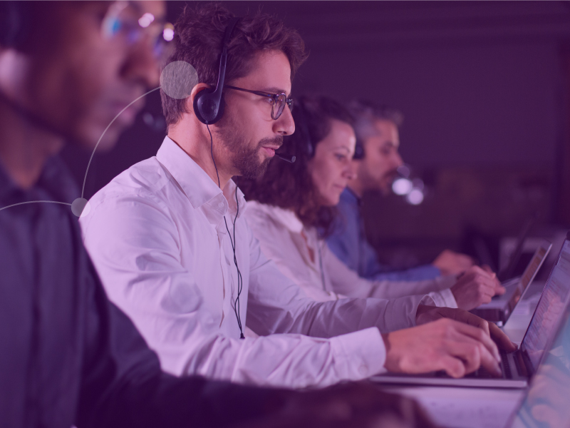 Homem usando um headset sorri com a mão no queixo olhando para o computador.